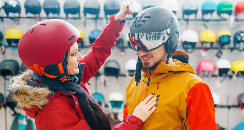 Two people in a store, one in a red helmet and jacket adjusting the helmet of the other, who is wearing a yellow and red jacket. They stand in front of a wall of various helmets, discussing their options for an upcoming skiing trip.