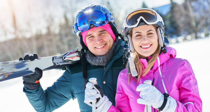 A man and a woman, both wearing ski gear and helmets, smile at the camera. The man holds skis over his shoulder, and the woman holds ski poles. Snowy landscape and trees are visible in the background, capturing their skiing adventure.