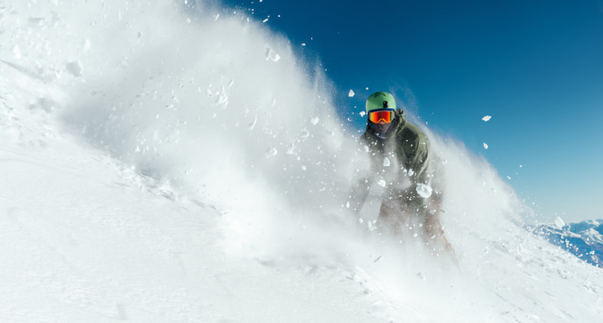 A snowboarder in a helmet and goggles rides through powder snow on a sunny skiing day.