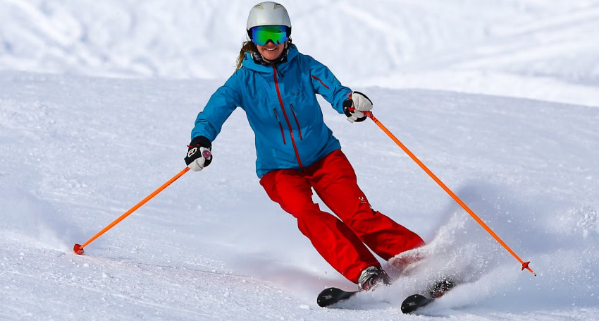 A person in a blue jacket and red pants is skiing downhill on snow, holding orange ski poles, with a white helmet and green-tinted goggles.