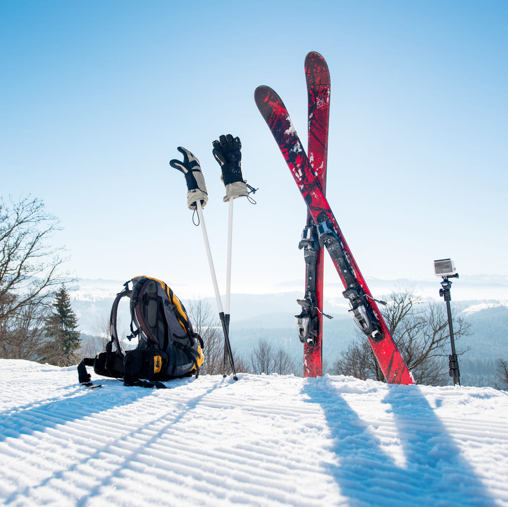 A pair of red skis, ski poles with gloves, a backpack, and a camera are placed on a snowy slope with trees and mountains in the background on a sunny day.