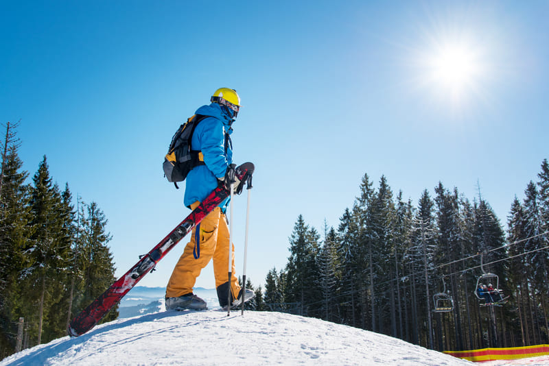 Person in colorful winter gear holding a snowboard and skis stands on a snowy hilltop, looking at distant mountains, with a sunlit sky and pine trees in the background.