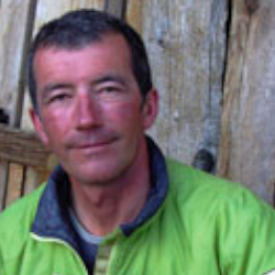 A middle-aged man with short dark hair and a slight smile, wearing a green jacket, stands in front of a wooden background.