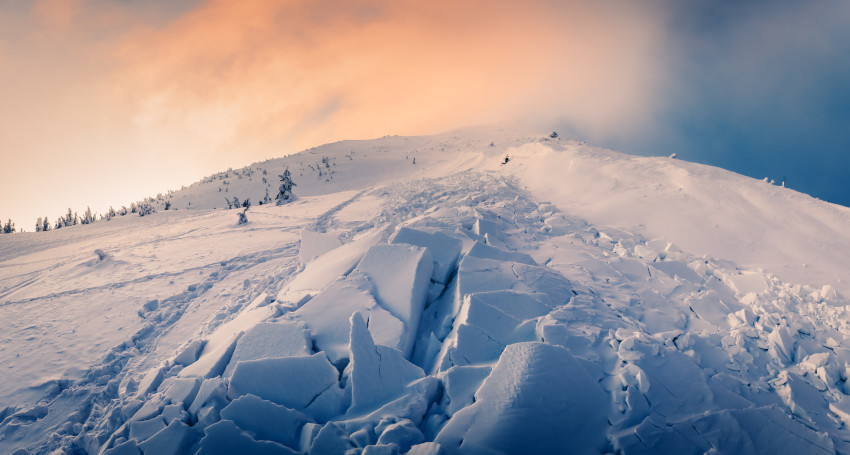 Panoramic view of a snow-covered mountainside, perfect for skiing, with cracked patches of snow and ice under a sky transitioning from pale yellow to blue, and a few scattered trees visible. Remember to wear your helmet as you navigate this stunning winter landscape.