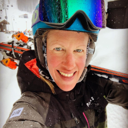 A person in ski gear and reflective goggles holds skis over their shoulder, standing in a snowy landscape.