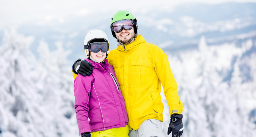 Two people in colorful ski outfits, one wearing a purple jacket and the other a yellow jacket, stand close together against a snowy, mountainous backdrop, their helmets adding an extra layer of safety for their exhilarating skiing adventure.