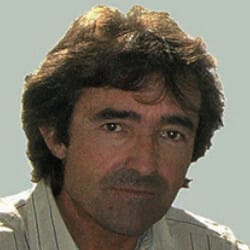 A man with wavy dark hair wearing a light-colored shirt looks directly at the camera against a plain background.
