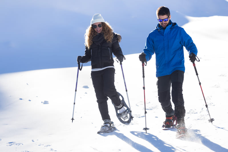 Two people in winter gear walk on a snowy landscape with poles, one in snowshoes and the other in boots. The scene is bright and sunny.