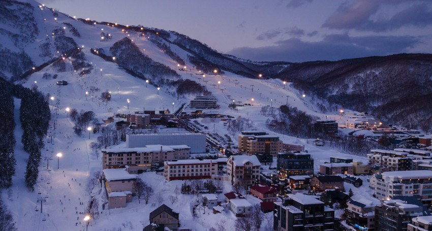 Aerial view of a snow-covered Hanazono Niseko ski resort at dusk, with lit-up slopes, buildings, and surrounding mountains.