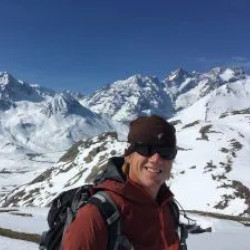 Ian Huyton wearing a red jacket, sunglasses, and a beanie stands in a snow-covered mountainous landscape on a sunny day.