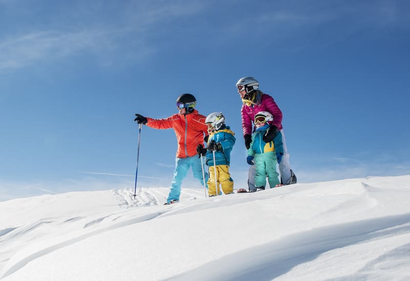 Four people dressed in colorful ski gear stand on a snowy slope with skis and poles, against a clear blue sky.
