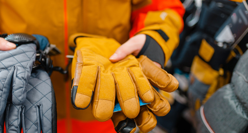 A person in an orange jacket holds tan leather gloves in one hand and gray quilted gloves in the other, demonstrating the importance of leather ski glove care.