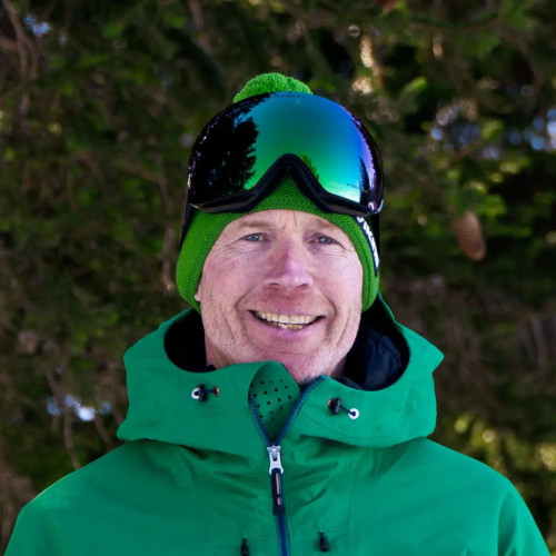 A person in green ski gear, including a jacket, beanie, and reflective goggles, smiles at the camera with a background of conifer trees.