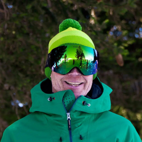A person wearing a green jacket, a green and yellow beanie, and reflective ski goggles smiles while standing outdoors with coniferous trees in the background.