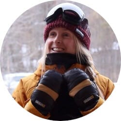 A person wearing a red beanie, ski goggles, and winter gloves smiles while dressed in a brown winter jacket.