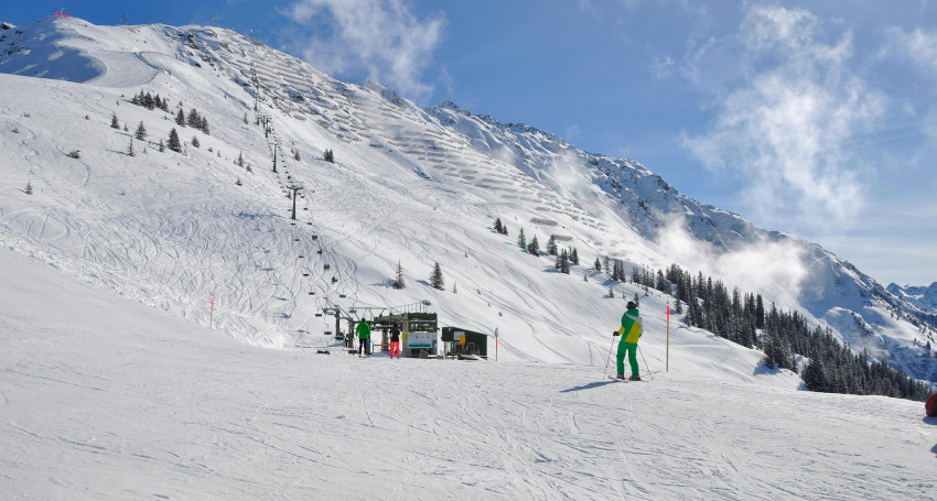 Skiers pause near a chairlift station on a snowy mountain in Mount Baker, Washington. A slope under a clear blue sky with scattered clouds. The mountainside and forested areas are covered in snow.
