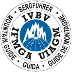 Circular logo for the International Federation of Mountain Guides Associations (IFMGA), featuring a mountainous graphic in the center and multilingual text around the border.