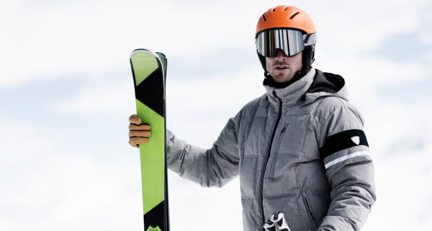 A person wearing a gray jacket and orange helmet stands holding skis in a snowy environment, pondering the question: Can you ski in glasses?