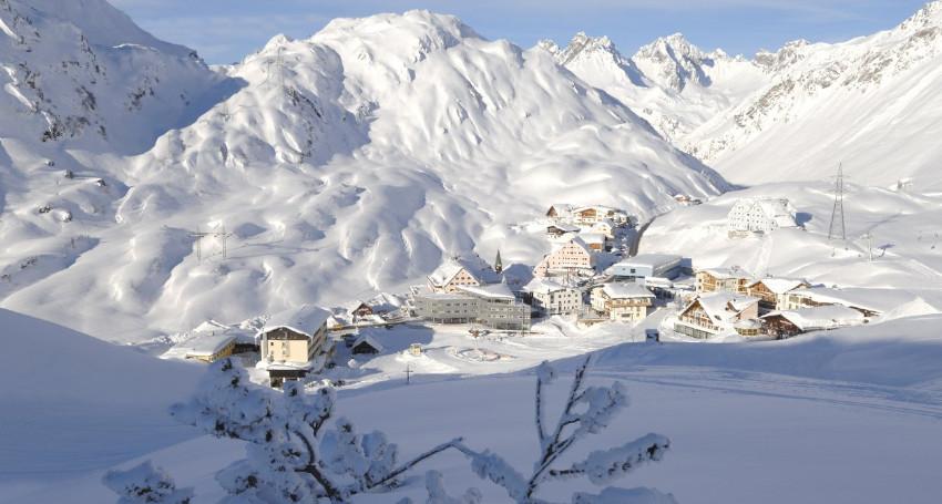 A snow-covered mountain village at St Anton nestled in a valley with scattered buildings and rugged peaks in the background.