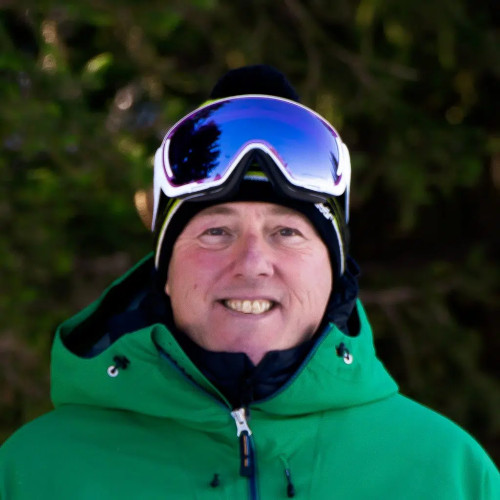 A person in a green jacket and ski goggles smiles outdoors with a background of trees.