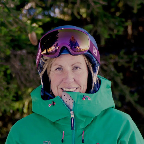 A person dressed in a green jacket and reflective ski goggles smiles outdoors with trees in the background.