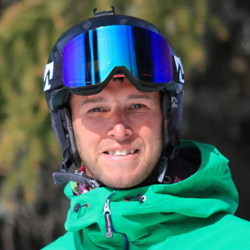 A person wearing a green jacket and a ski helmet with mirrored goggles smiles at the camera.