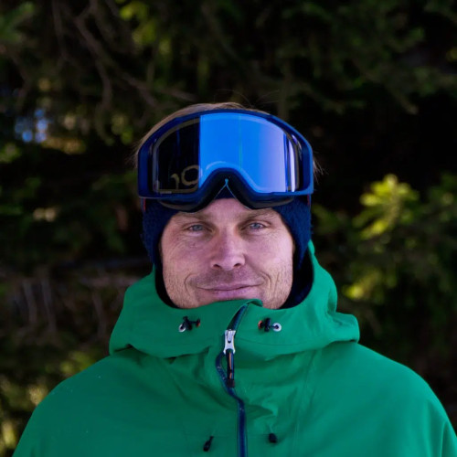 A person in a green jacket and blue ski goggles stands outdoors with trees in the background.
