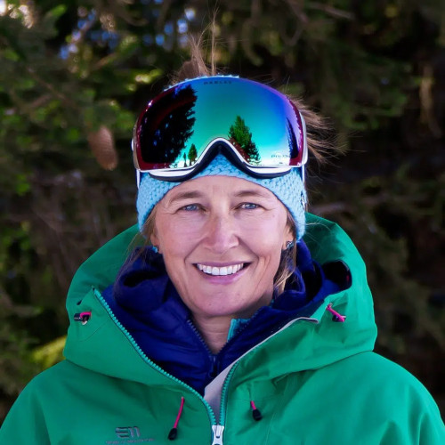 A person in winter gear, including reflective ski goggles and a green jacket, is smiling outdoors in a snowy, forested area.