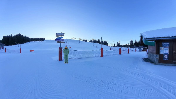 A person in a green suit stands near signposts on a snowy ski slope with nets and barriers. A wooden building with a green roof is on the right. Trees and ski lifts are visible in the background.