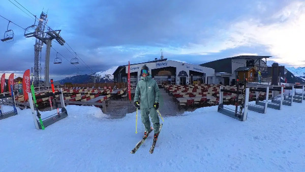 A skier stands with skis and poles on a snowy slope in front of a mountain lodge with empty outdoor seating, under an overcast sky. Ski lifts and mountains are visible in the background.