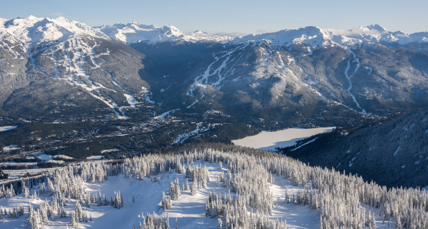 Aerial view of snow-covered mountains in Whistler Blackcomb, Canada with a frozen lake, and dense forests under a clear sky.
