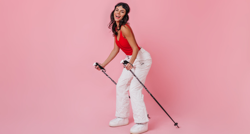 A woman in a red top and white pants poses with ski poles, also known as ski sticks, against a pink background.